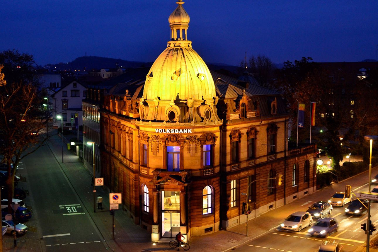 Die kunstvolle Fassade des Volksbank-Gebäudes bei Nacht, beleuchtet von warmen goldenen Lichtern.