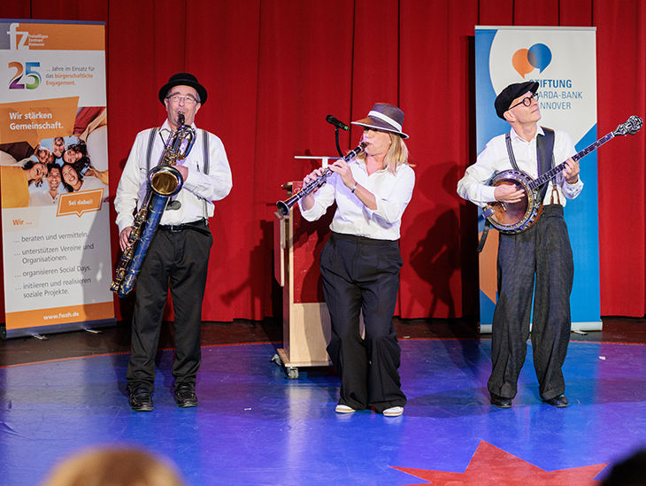 Drei Musiker spielen auf einer Bühne vor rotem Vorhang Saxophon, Klarinette und Banjo.