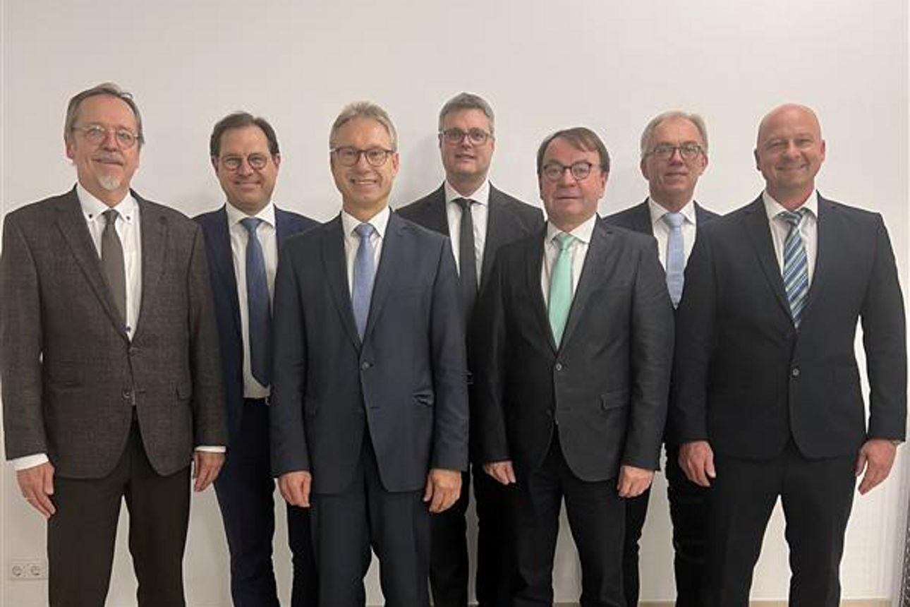 Sieben Männer in Anzug stehen als Stiftungsbeirat vor einer Wand