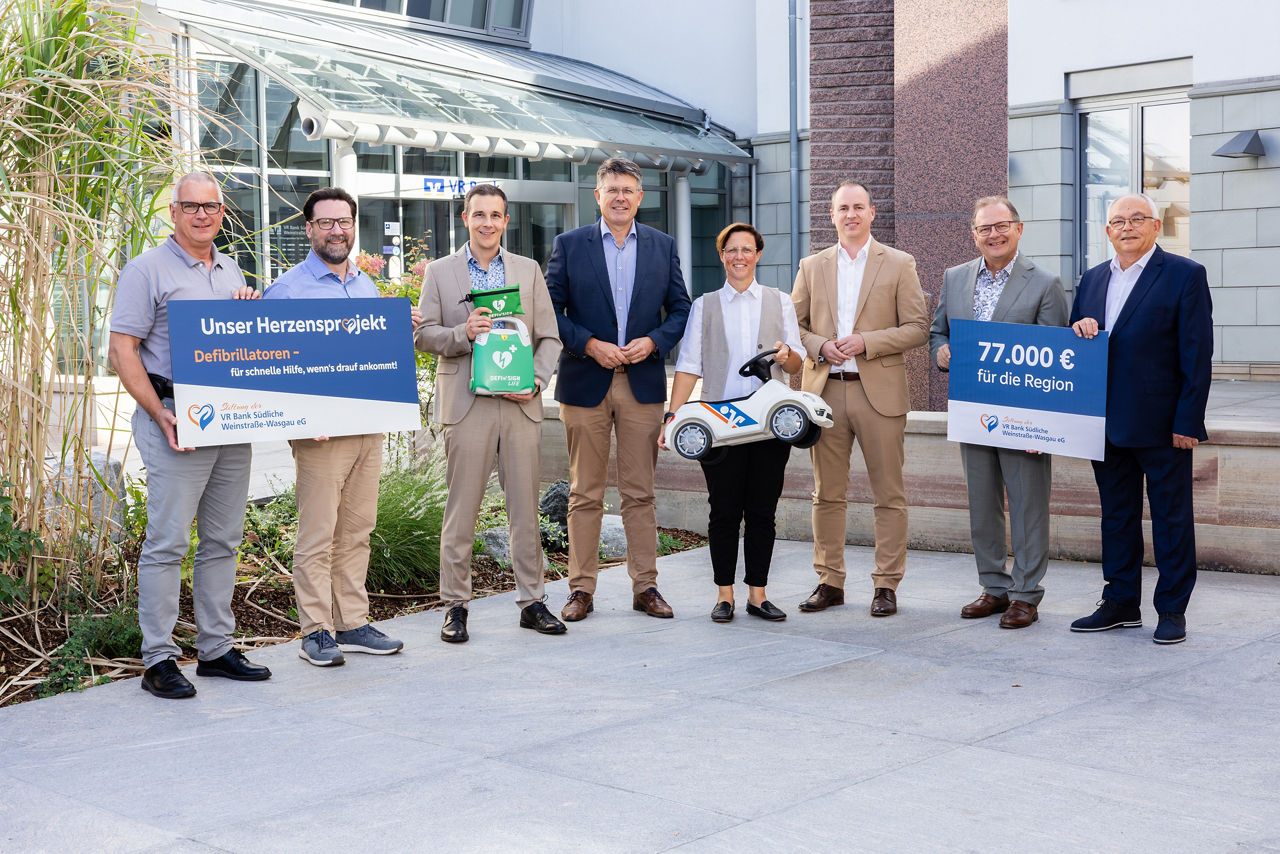 Gruppenfoto vor einer Bankfiliale mit Informationstafeln zum Defibrillator-Projekt und regionaler Förderung.
