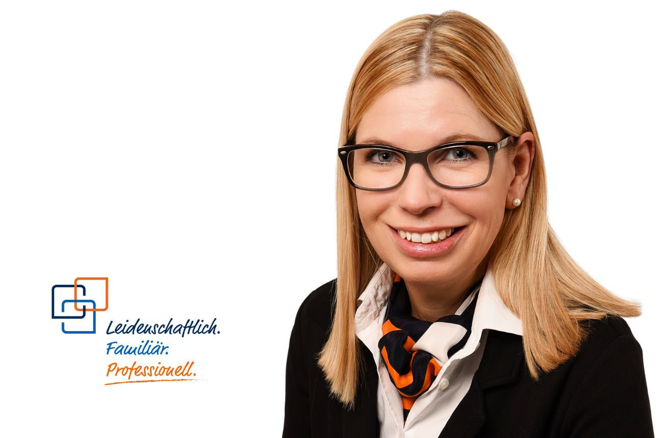 Anja Stillecke, lächelnd, mit Brille, blonden Haaren und einem gemusterten Schal in Orange, Weiß und Blau.