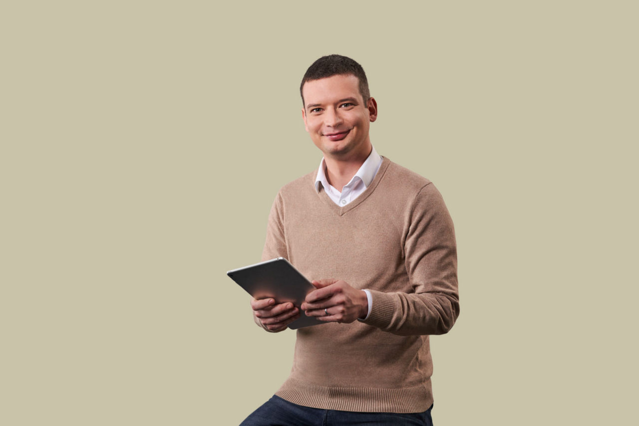 Mitarbeiter sitzt mit Tablet in der Hand vor beigem Hintergrund und schaut in die Kamera