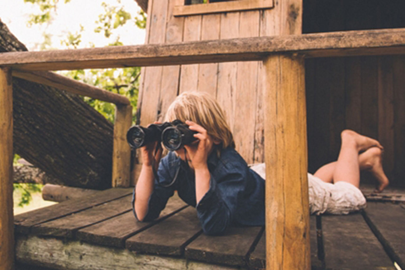 Kind liegt auf der Veranda seines Baumhauses und schaut durch ein Fernglas.