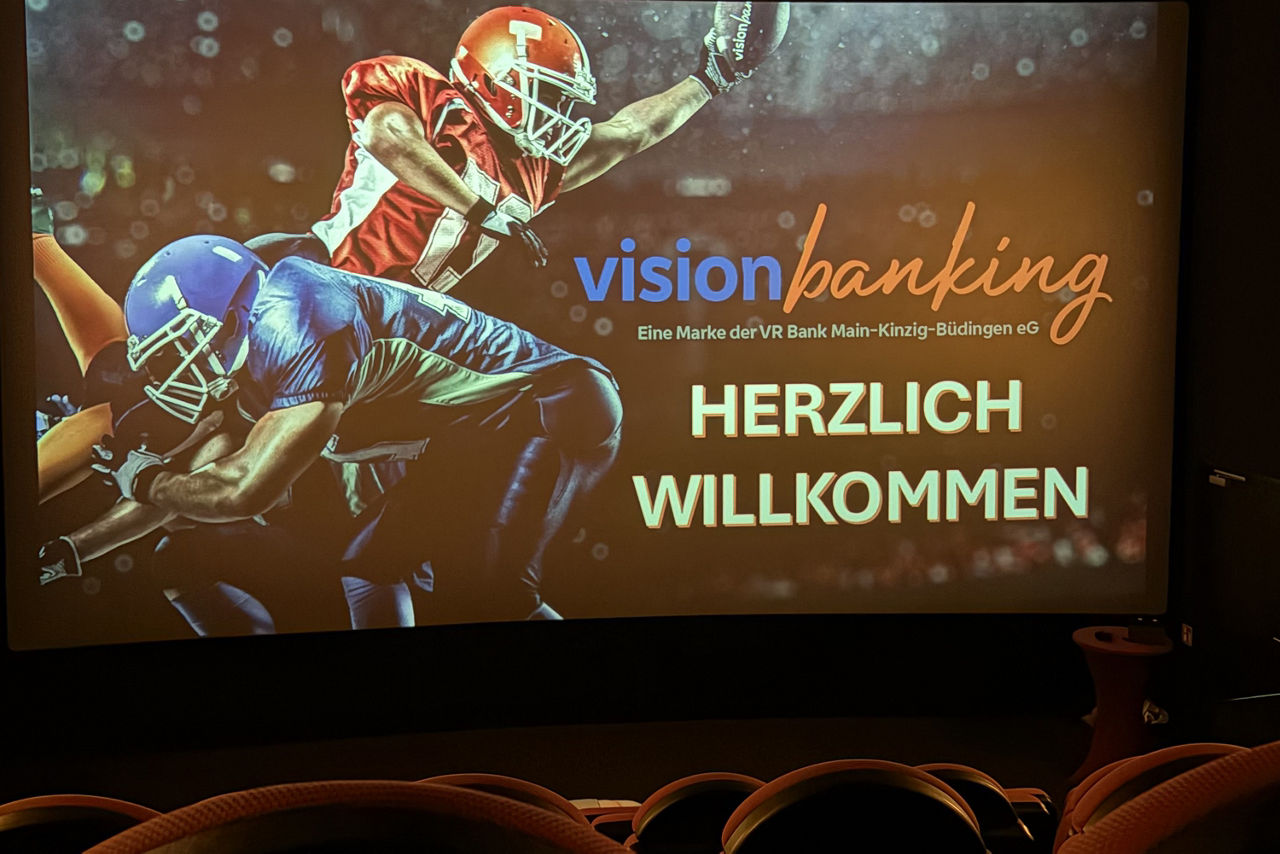 Das Bild zeigt zwei Footballspieler auf einer Kinoleinwand, mit der Aufschrift Herzlich Willkommen. Im Vordergrund sind Kinosessel zu sehen