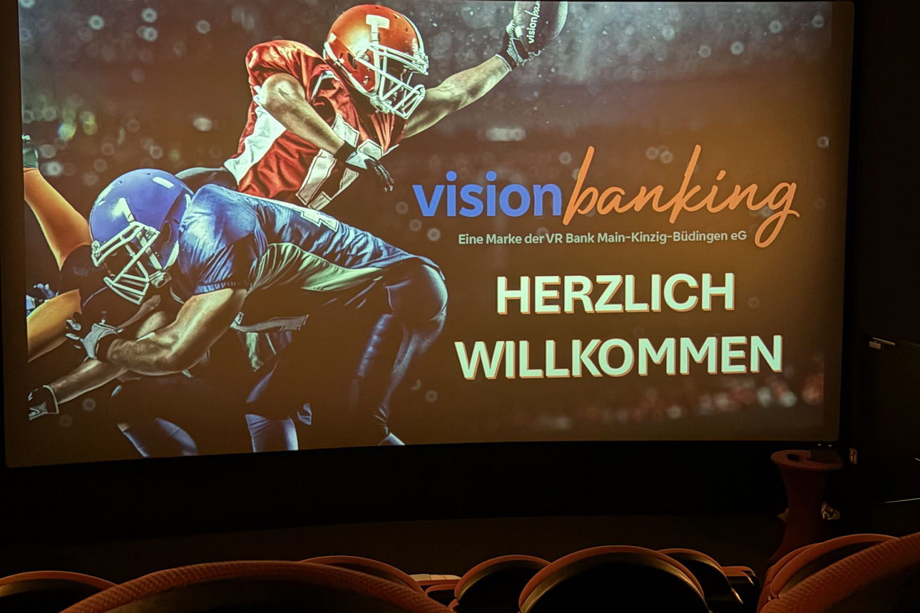 Das Bild zeigt zwei Footballspieler auf einer Kinoleinwand, mit der Aufschrift Herzlich Willkommen. Im Vordergrund sind Kinosessel zu sehen