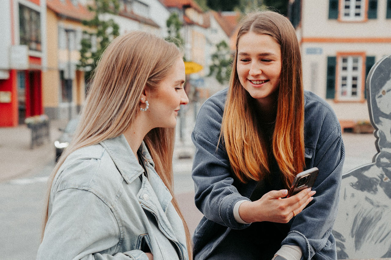 Zwei Personen sitzen in der Stadt, eine zeigt der anderen etwas auf dem Smartphone