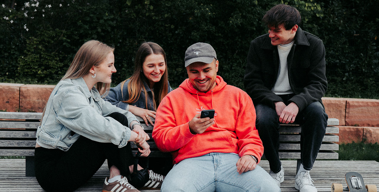 Vier Personen sitzen draußen auf einer Bank, eine trägt einen orangen Hoodie und hält ein Smartphone