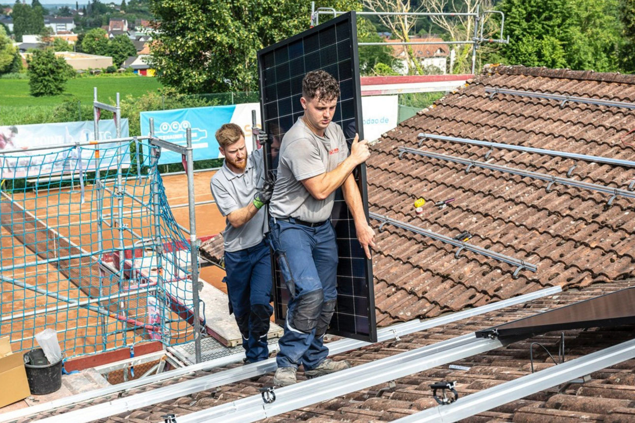 Auf dem Bild sieht man zwei Männer, die ein Modul einer Photovoltaikanlage auf einem Dach tragen
