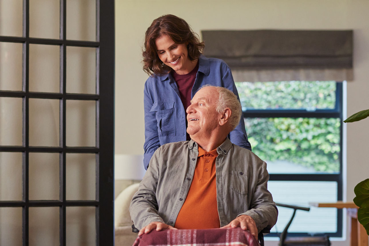 Eine Frau fährt einen Mann auf einem Rollstuhl und sie lächeln sich an.