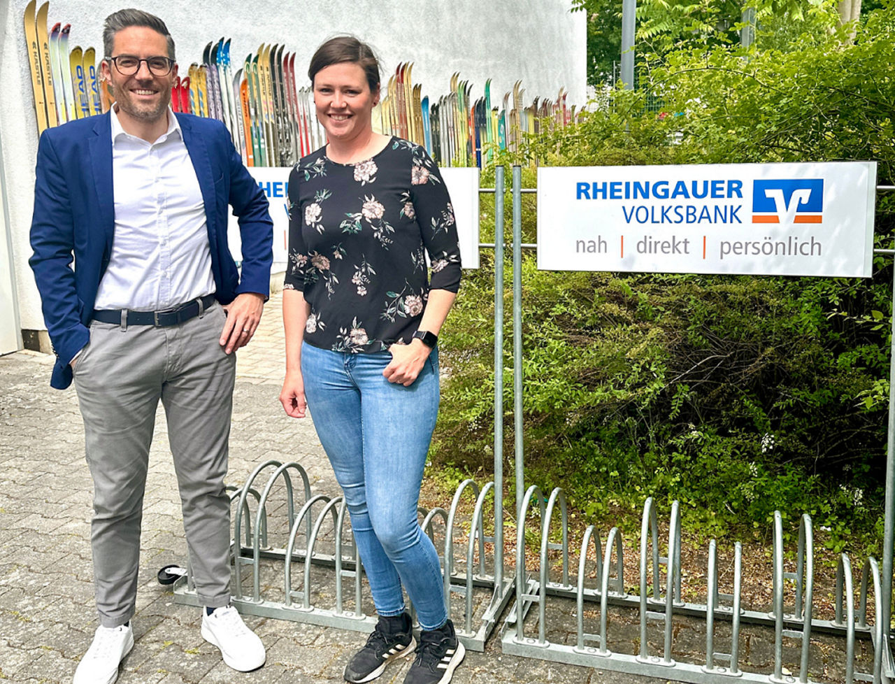 Zwei Personen stehen vor einem Fahrradständer mit einem Schild der Rheingauer Volksbank. Im Hintergrund sind mehrere Skier aufgestellt.