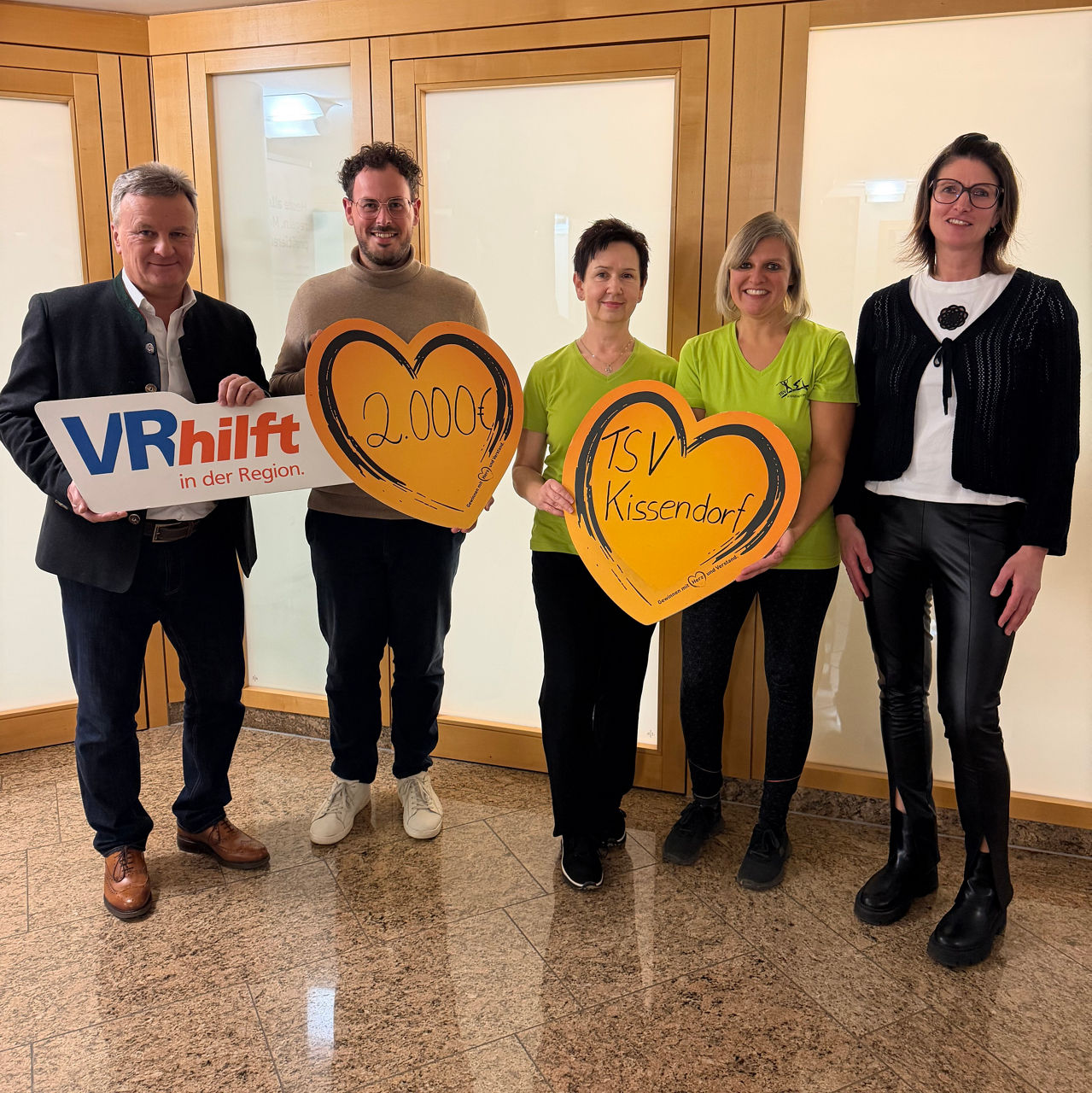 Fünf Personen mit Herzschildern und „VR hilft“-Logo, gemeinsame Spendenübergabe in Innenraum