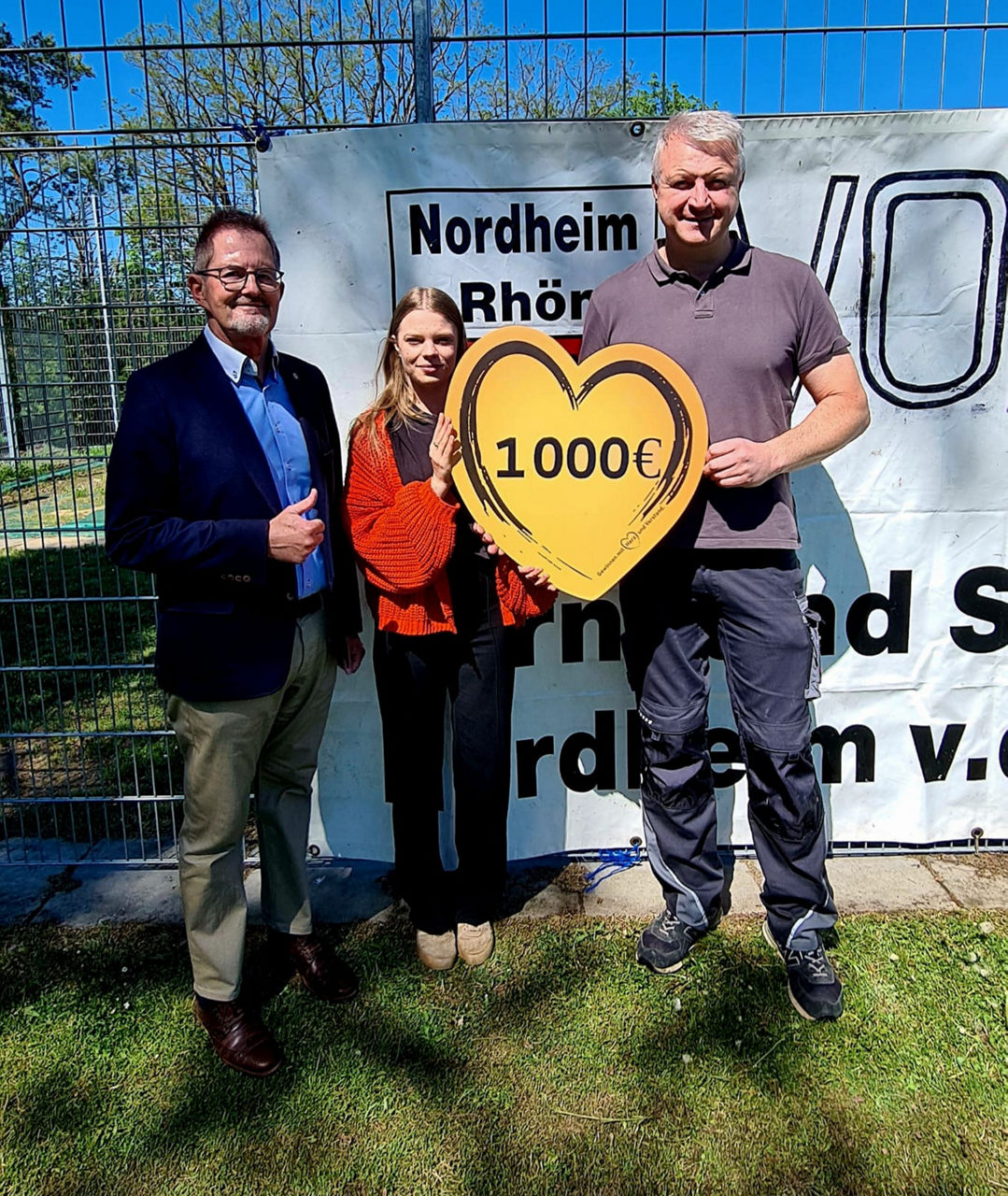 Drei Personen stehen vor einemben steht ein weiterer Mann mit einem Daumen-hoch-Zeich Vereinsbanner des TSV Nordheim. In der Mitte hält eine Frau gemeinsam mit einem Mann ein großes Herzschild mit der Aufschrift „1.000 €“. Links daneben. Die Spende stammt von der VR-Bank Main-Rhön zur Unterstützung des Vereins.