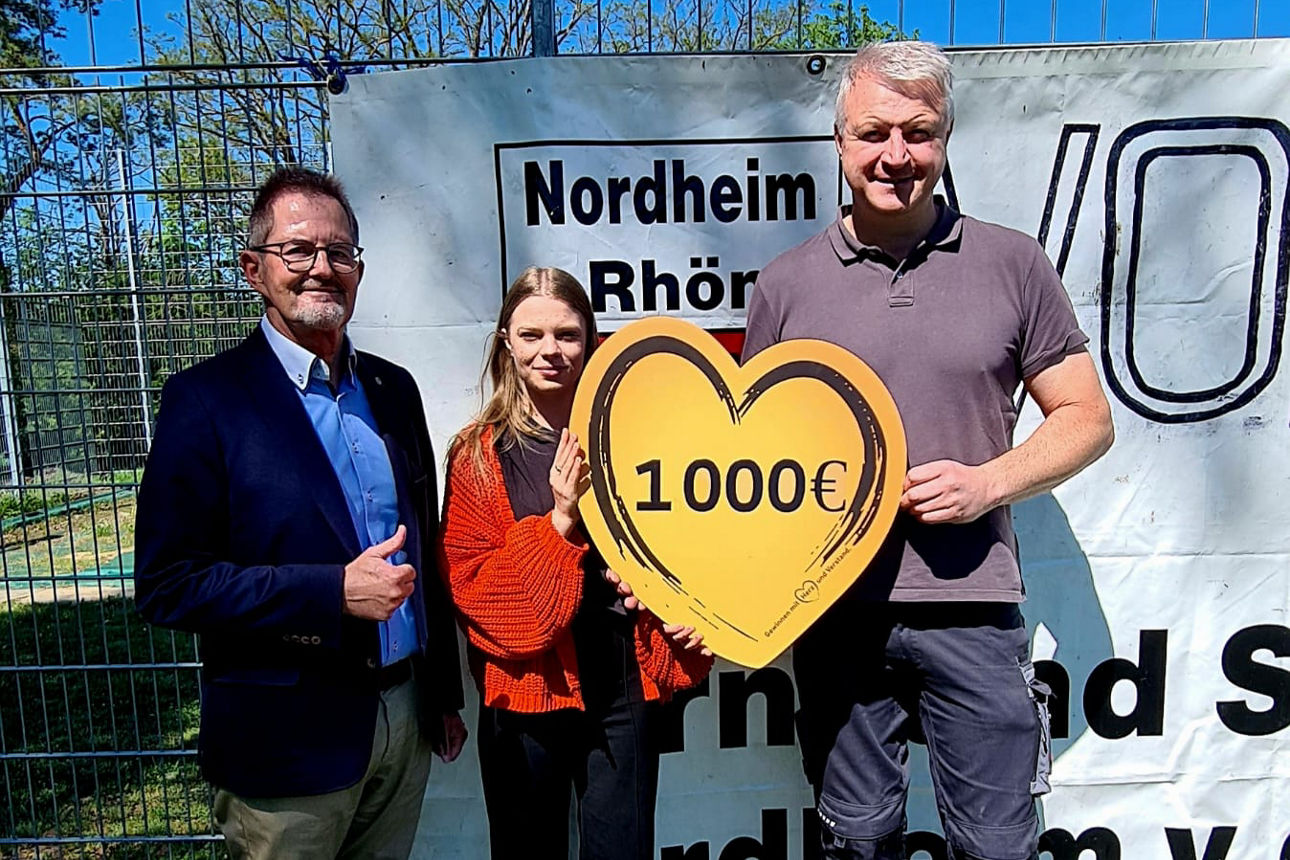 Drei Personen stehen vor einemben steht ein weiterer Mann mit einem Daumen-hoch-Zeich Vereinsbanner des TSV Nordheim. In der Mitte hält eine Frau gemeinsam mit einem Mann ein großes Herzschild mit der Aufschrift „1.000 €“. Links daneben. Die Spende stammt von der VR-Bank Main-Rhön zur Unterstützung des Vereins.