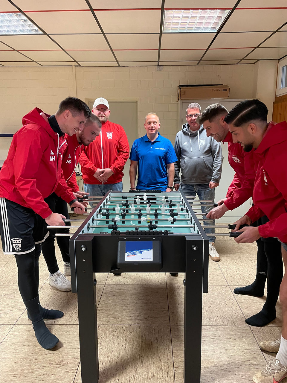 Eine Gruppe von Männern spielt Tischfußball in einem Raum mit gefliestem Boden und gefliester Decke