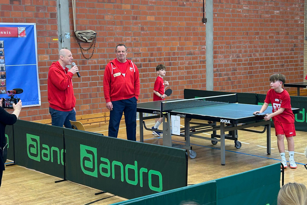 Man kann die Aufteilung in einer Sporthalle für mehrere Tischtennisplatten erkennen. Zwei Männer halten eine Ansprache.