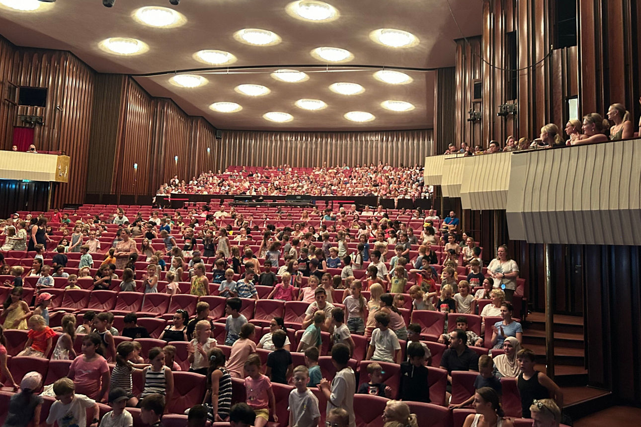 Theatersaal; im Publikum sitzen Kinder, die gemeinsam singen.