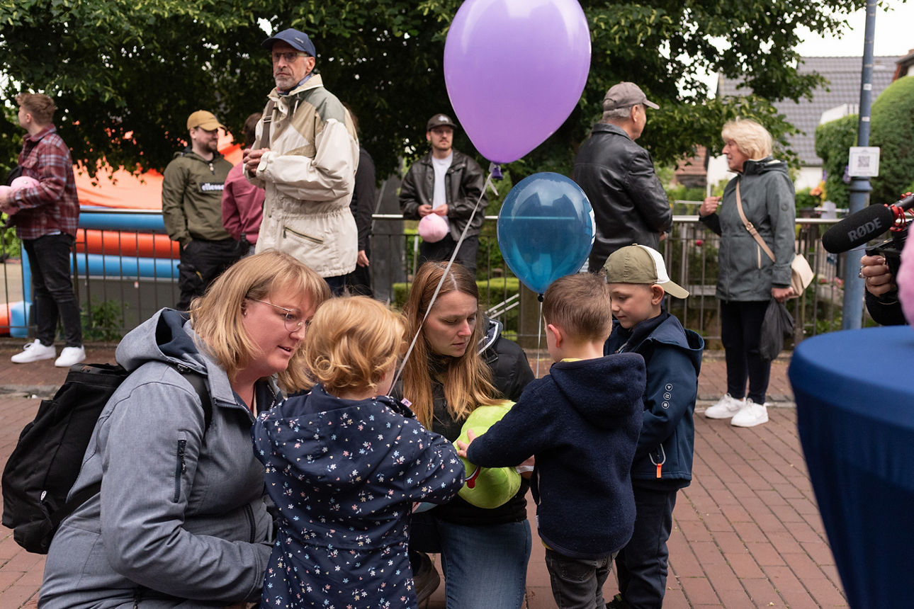 Drei Kinder und zwei knieende Frauen, ein Kind hält einen pinken Luftballon