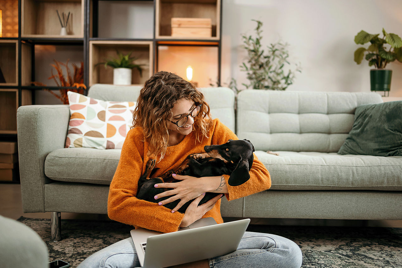 Frau sitz auf dem Bodem mit einem Laptop auf dem Schoß und einem Hund im Arm.
