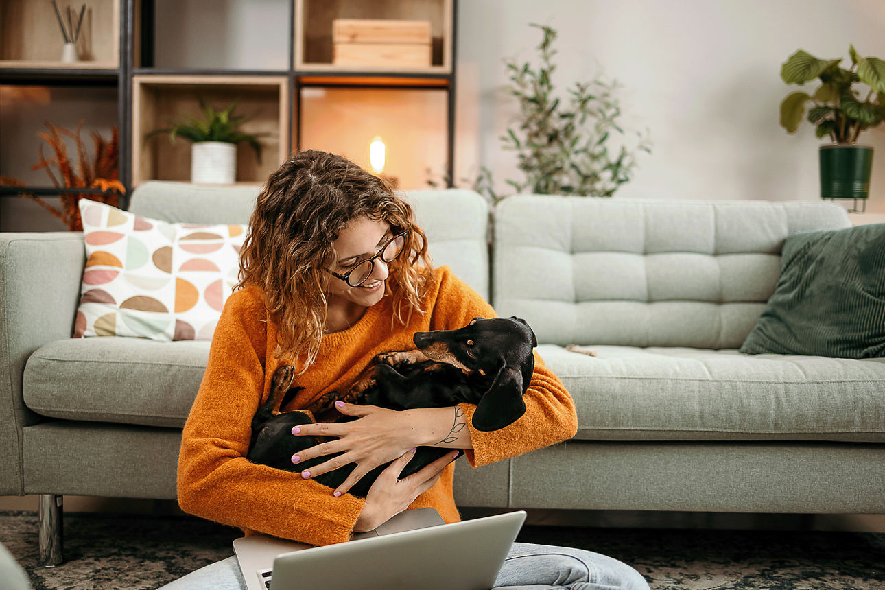 Frau sitz auf dem Bodem mit einem Laptop auf dem Schoß und einem Hund im Arm.