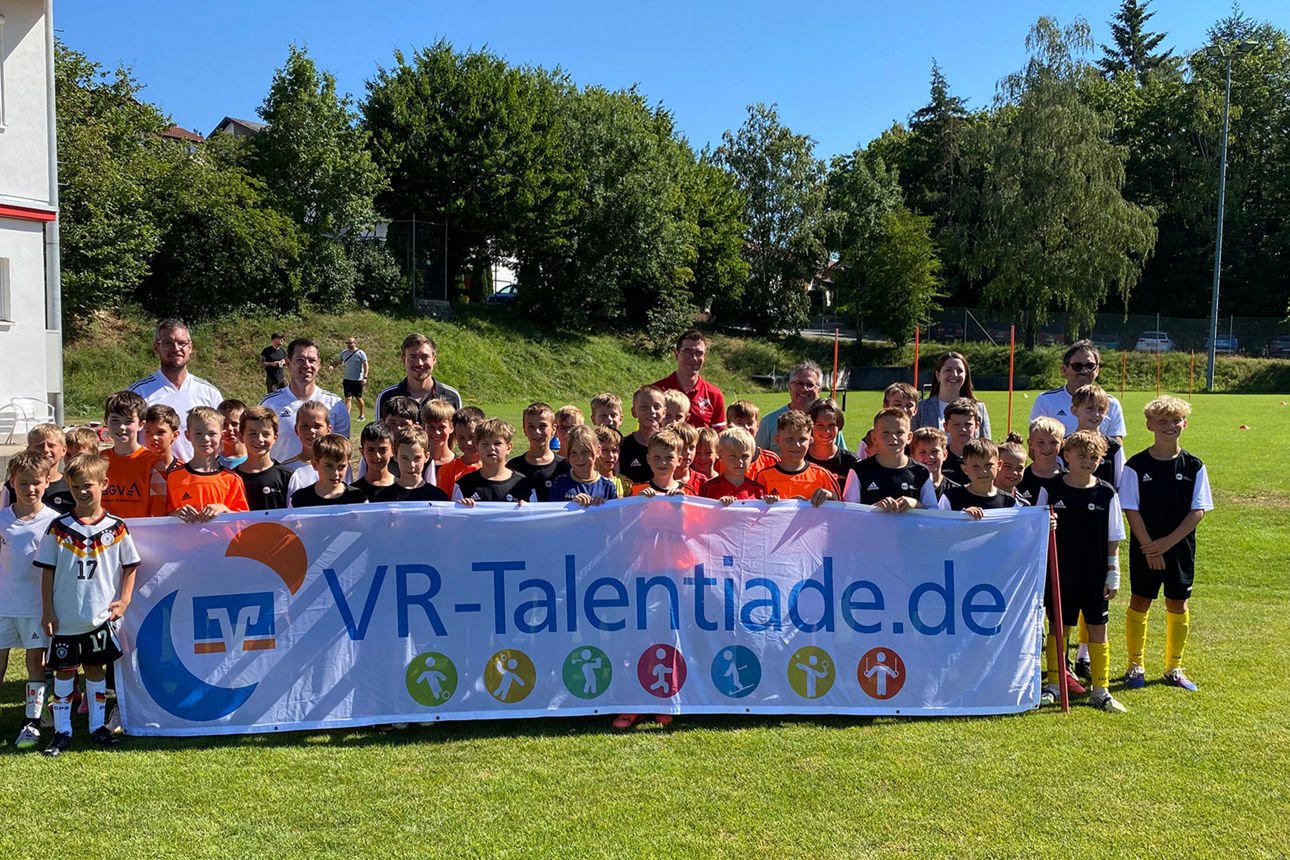 Kinder und Erwachsene stehen auf einem Fußballfeld und halten ein Banner mit der Aufschrift „VR-Talentiade.de“