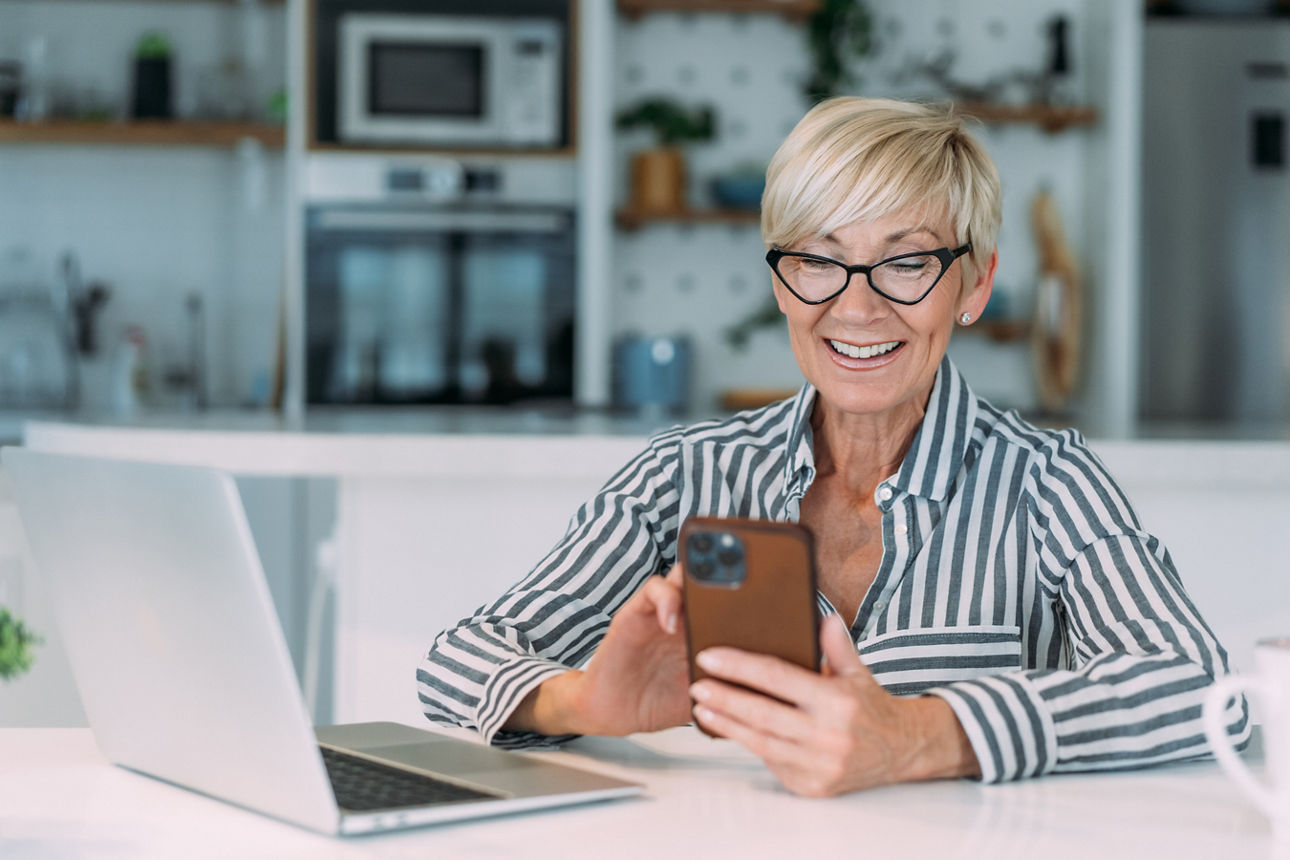 Eine lächelnde Frau mit Brille sitzt am Tisch, schaut auf ihr Smartphone und hat einen Laptop und eine Tasse vor sich.