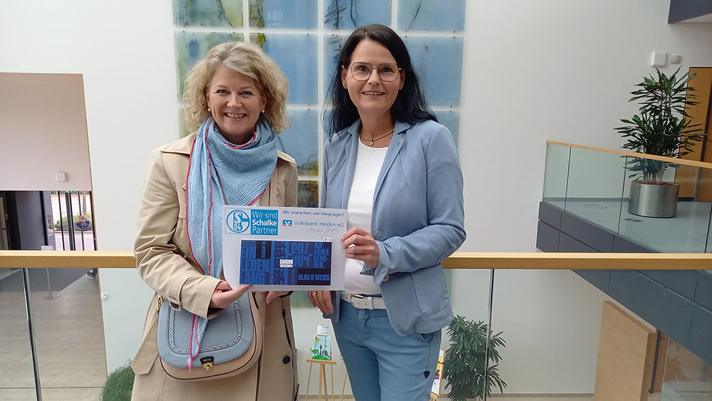Zwei Frauen stehen vor einem Gebäude und halten ein blaues Schild.