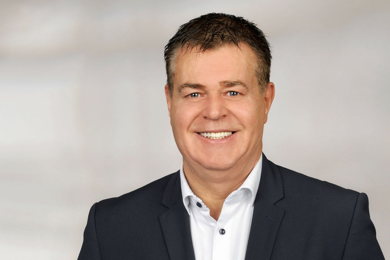 Mann mit kurzen dunklen Haaren trägt weißen Hemdkragen und dunkelblauen Anzug, vor unscharfem Hintergrund