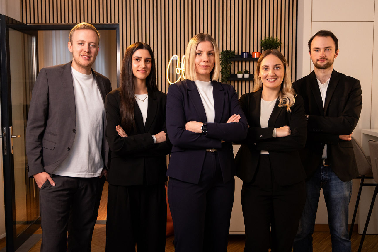 Fünf Personen in Businesskleidung stehen selbstbewusst nebeneinander in einem modern gestalteten Büro mit Holz- und Lichtelementen.