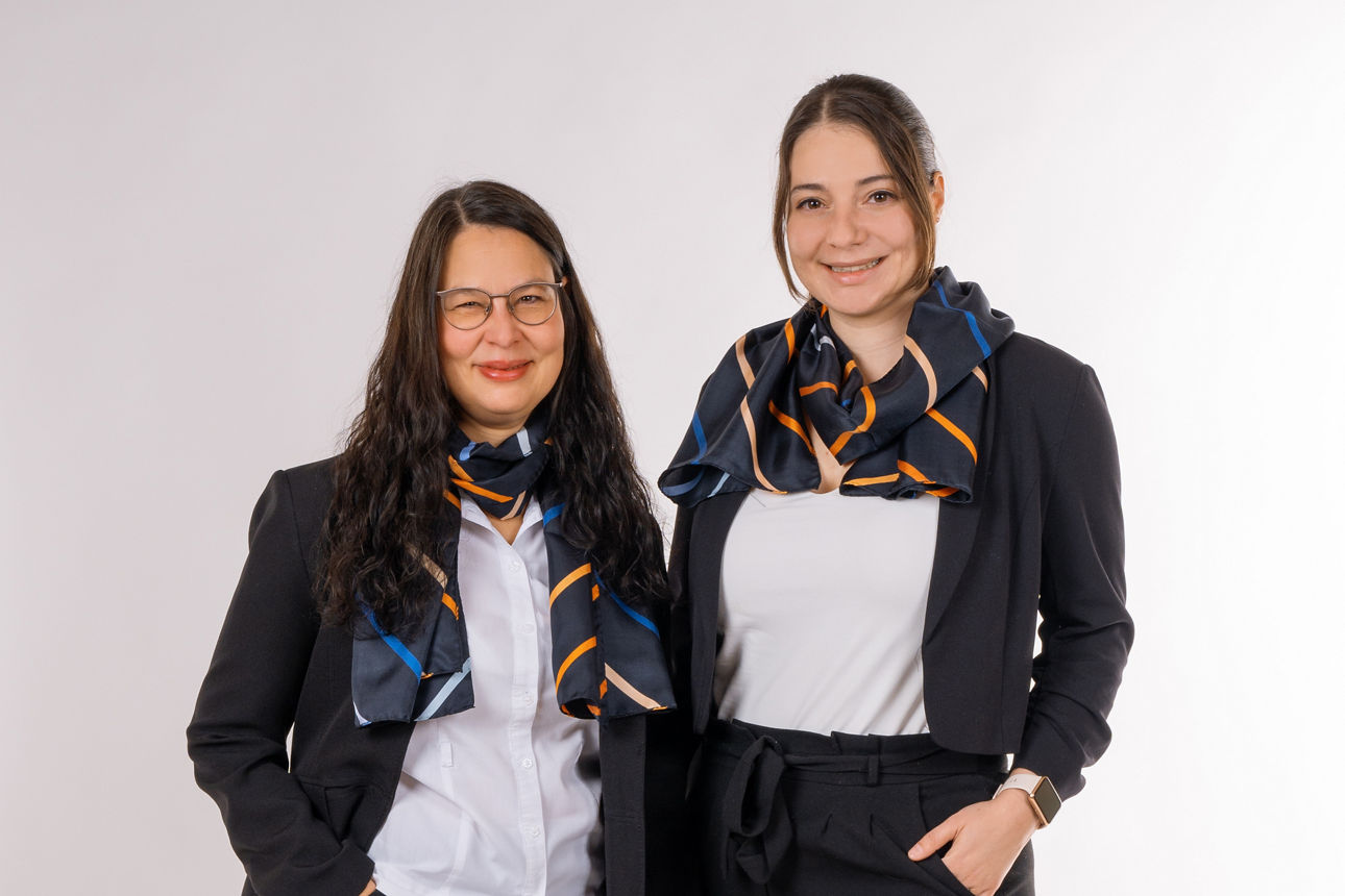 Zwei Frauen von der ZV-Beratung, in Geschäftskleidung mit Schals gekleidet, stehen nebeneinander und lächeln in die Kamera.