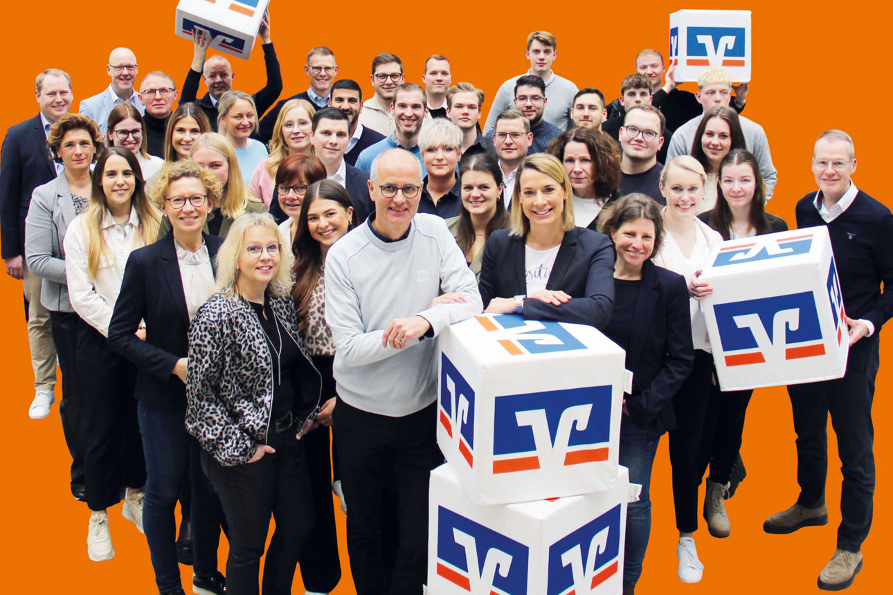 Team der Volksbank Baumberge eG mit Volksbank Würfeln vor orangem Hintergrund
