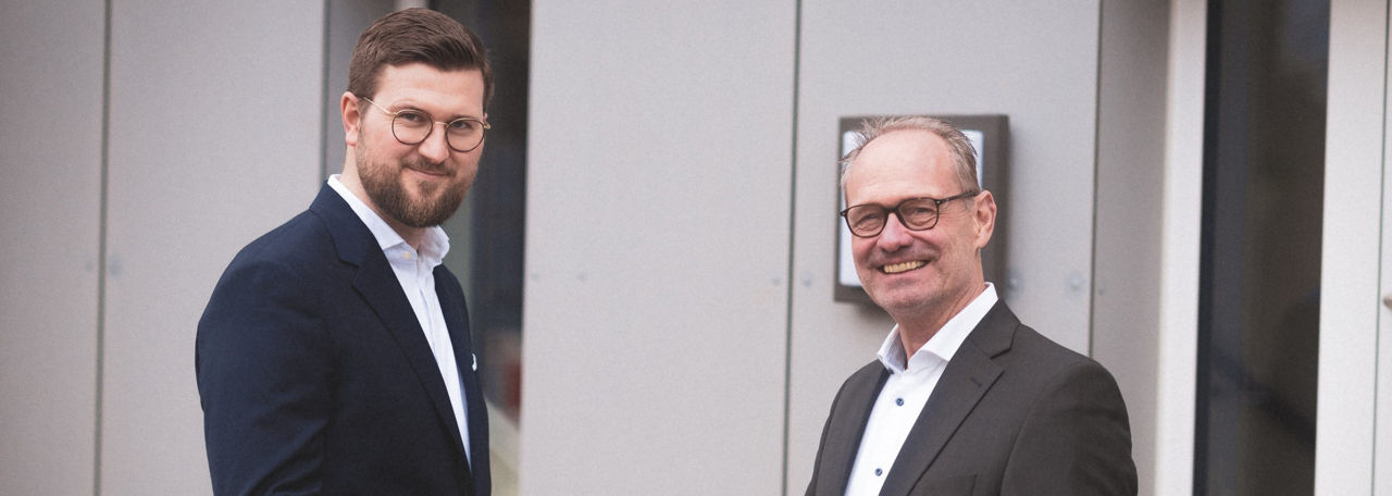 Zwei Männer in Anzügen und Brillen stehen nebeneinander und lächeln vor einem modernen Gebäude.