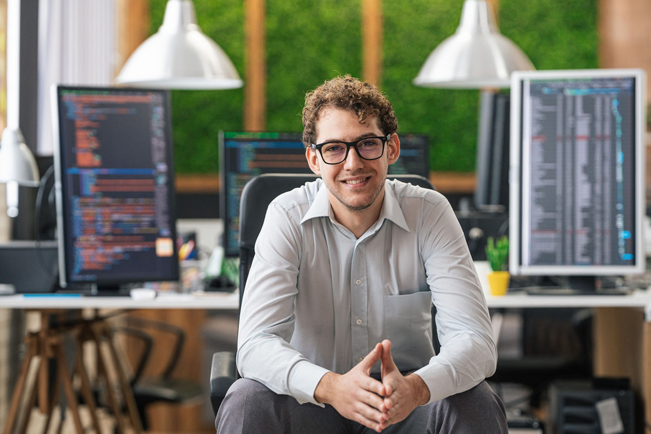 Portrait of smiling IT Programmer sitting near Desktop Computers. Male Specialist Creating Innovative Software. Engineer Developing App, Program, Video Game. Writing Code in Terminal