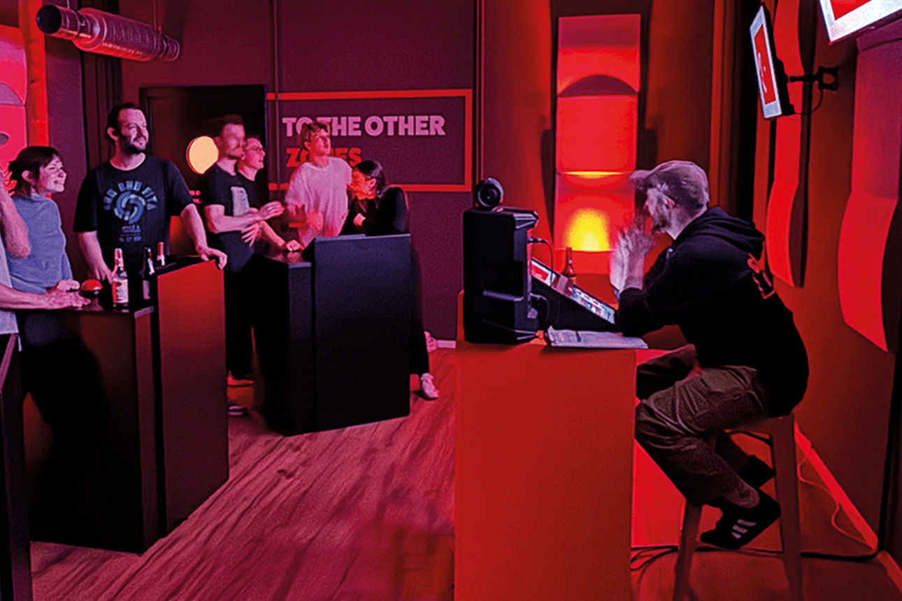 Sieben Menschen stehen an zwei Pulten; gegenüber sitzt ein Mann an einem Pult