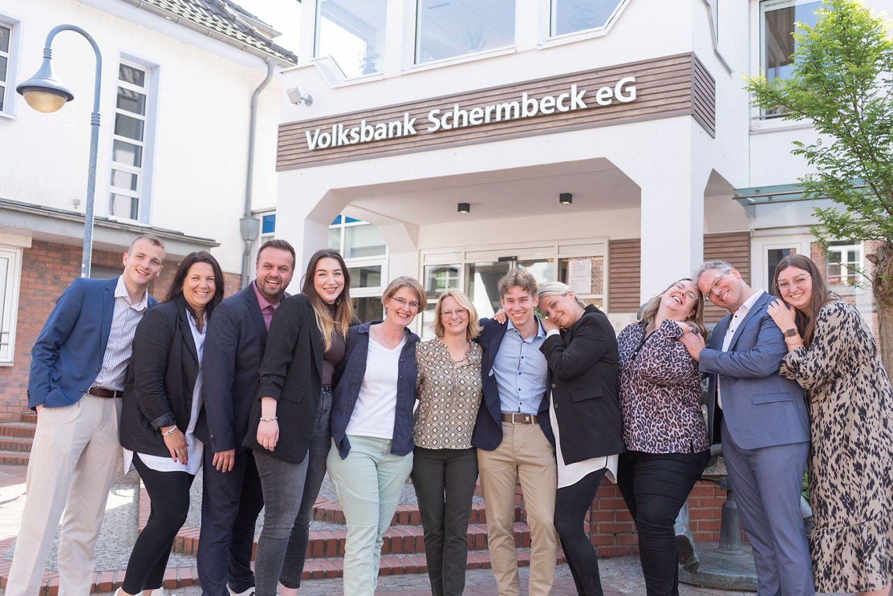 Mitarbeitende der Volksbank Schermbeck stehen lachend Arm in Arm vor der Bank.