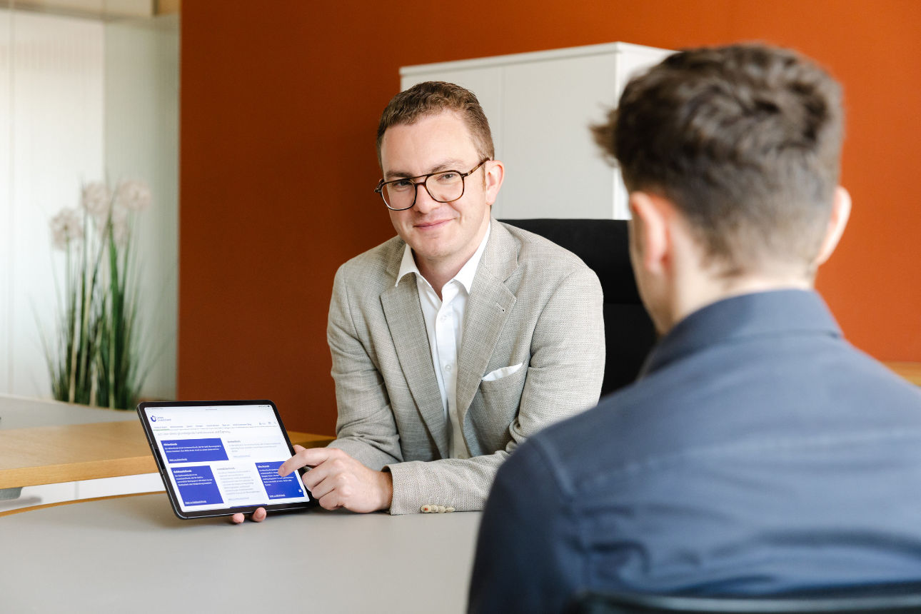 Zwei Personen in einem Büro, wobei eine Person auf einem Tablet Informationen präsentiert und diese der anderen erklärt.