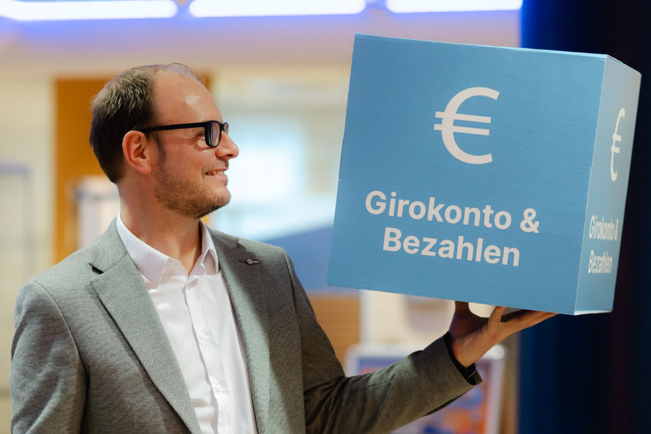 Eine Person in einem hellen Innenraum, die einen großen blauen Würfel in der Hand hält.