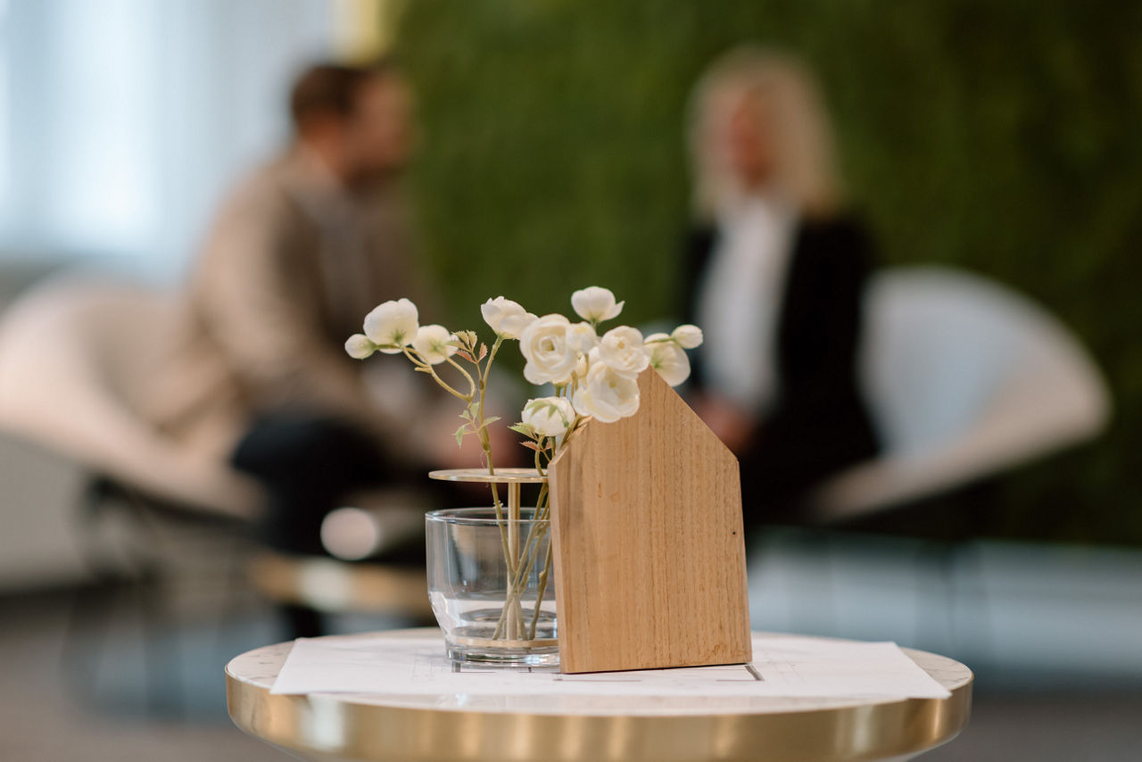 Im Vordergrund steht ein Holzhaus auf einem Tisch, im Hintergrund sitzen ein Mann und eine Frau.
