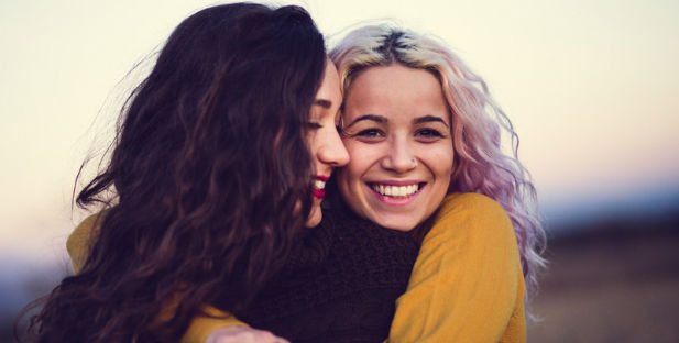 Zwei junge Frauen, eine mit langem, dunklem, lockigem Haar und die andere mit rosa Haaren, umarmen sich