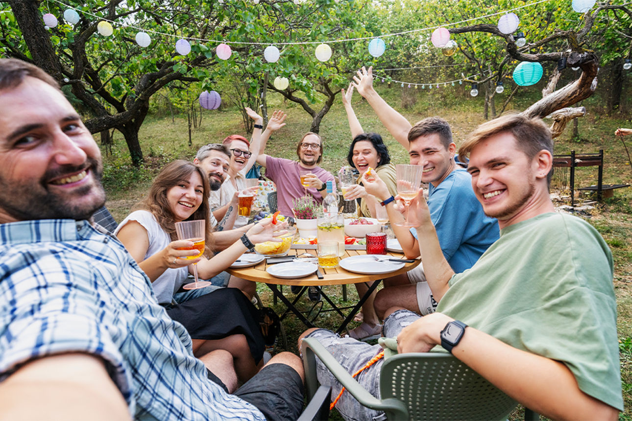 Freundesgruppe grillt im Garten, alle schauen fröhlich zur Kamera und prosten sich zu