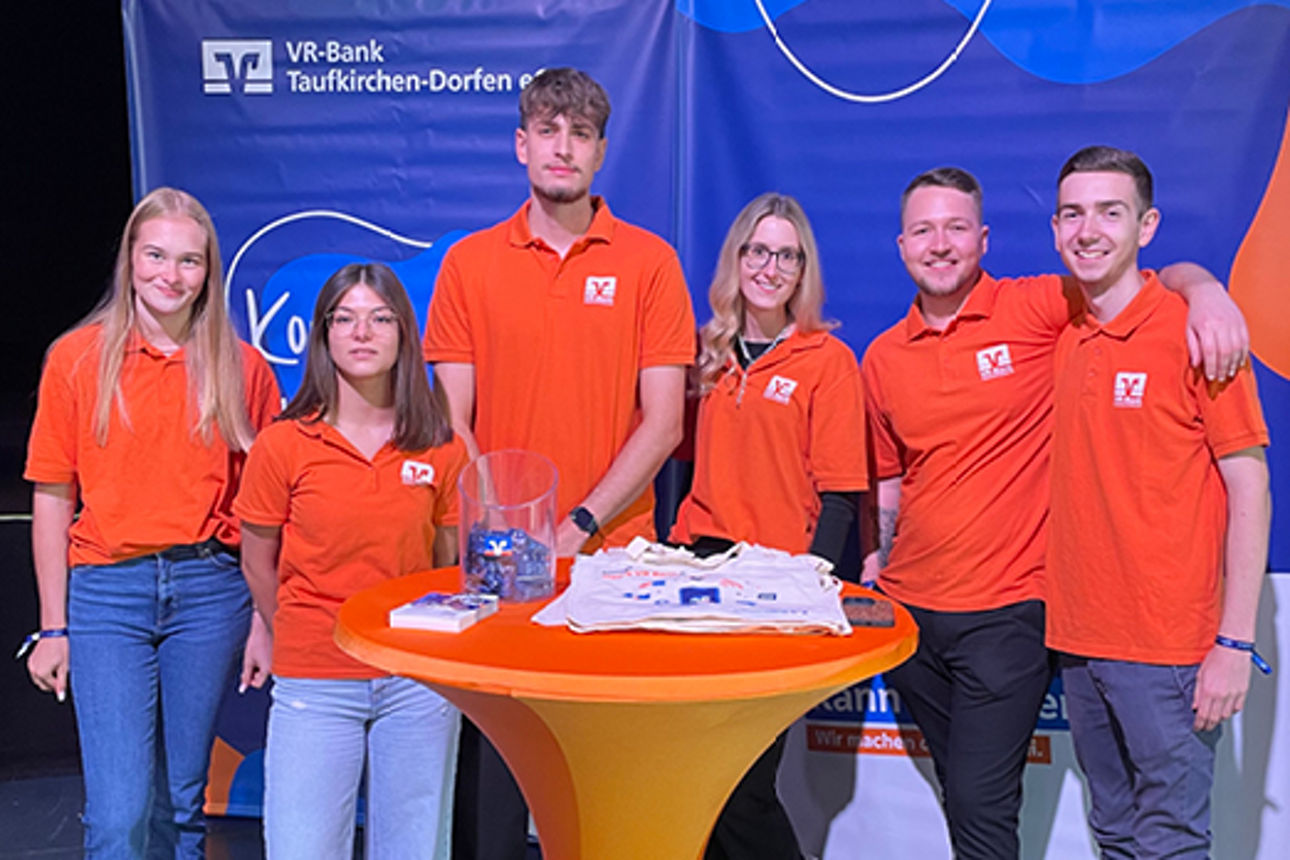 Sechs Kolleginnen und Kollegen stehen in orangen VR-Bank Shirts vor unserer Messewand.