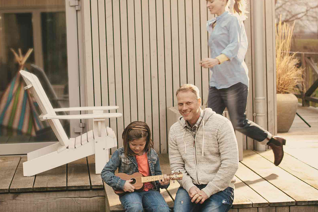 Vater sitzt mit seiner Tochter auf der Terrasse. Das Kind spielt Gitarre und im Hintergrund läuft die Mutter entlang.
