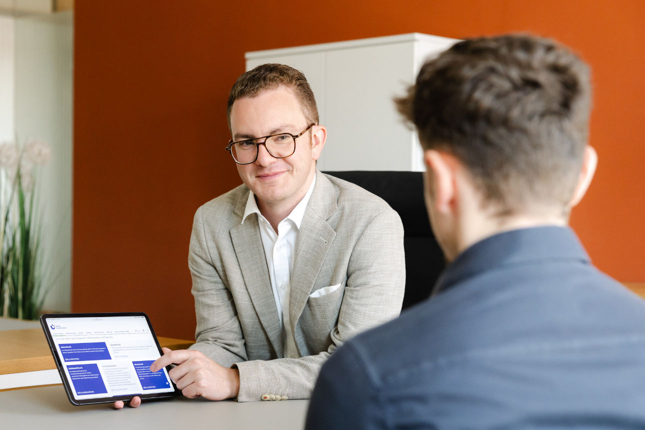 Zwei Personen in einem Büro, wobei eine Person auf einem Tablet Informationen präsentiert und diese der anderen erklärt.