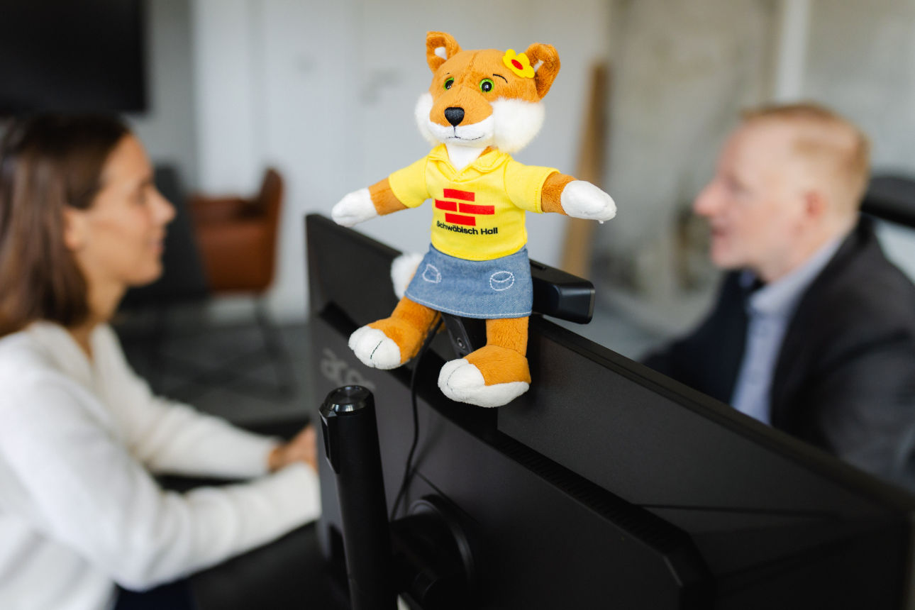 Ein Fuchs‑Stofftier mit gelbem T‑Shirt sitzt auf einem Monitor, während im Hintergrund zwei Personen arbeiten.
