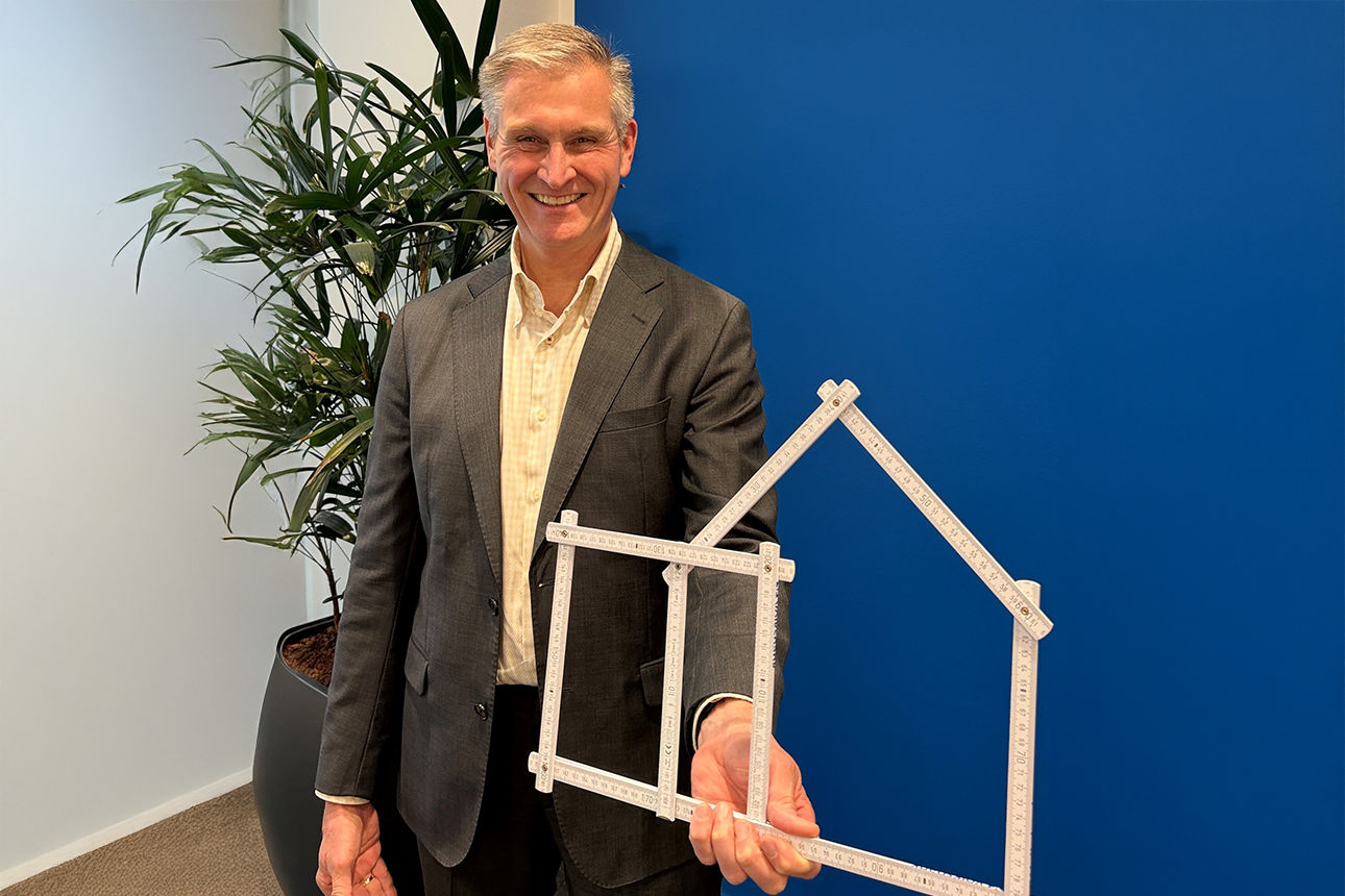 Mann steht vor einer blauen Wand mit einen Haus aus einem Zollstock in der Hand