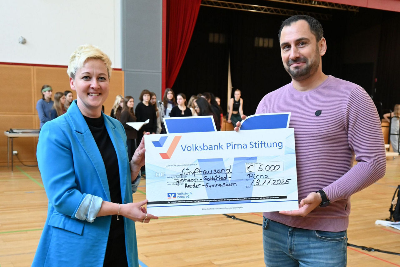 Blonde Frau in blauem Blazer übergibt einen Scheck über 5.000 Euro an einen Herren, beide stehen in einer Mehrzweckhalle