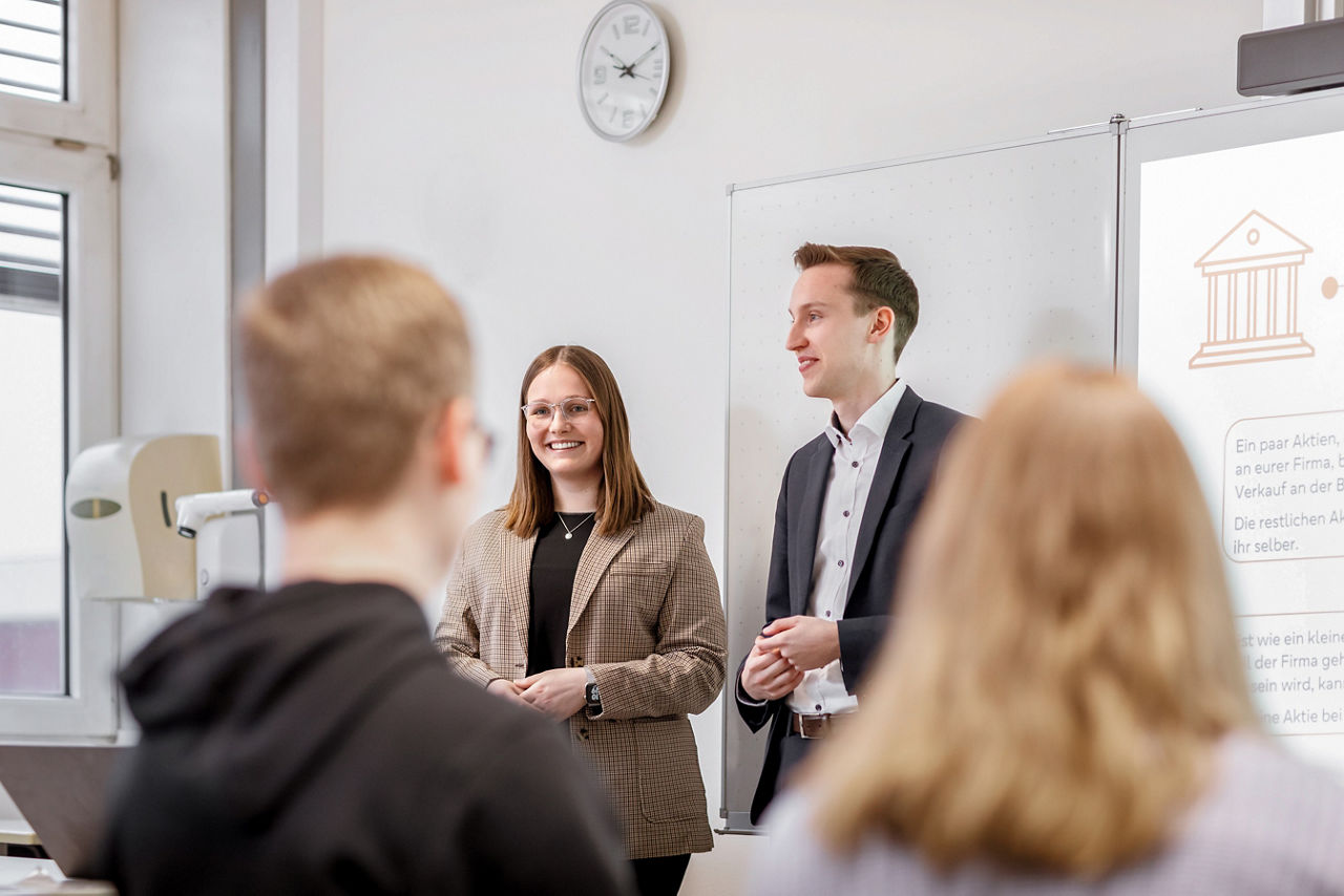Zwei Personen stehen vor einer Schulklasse und stellen etwas vor