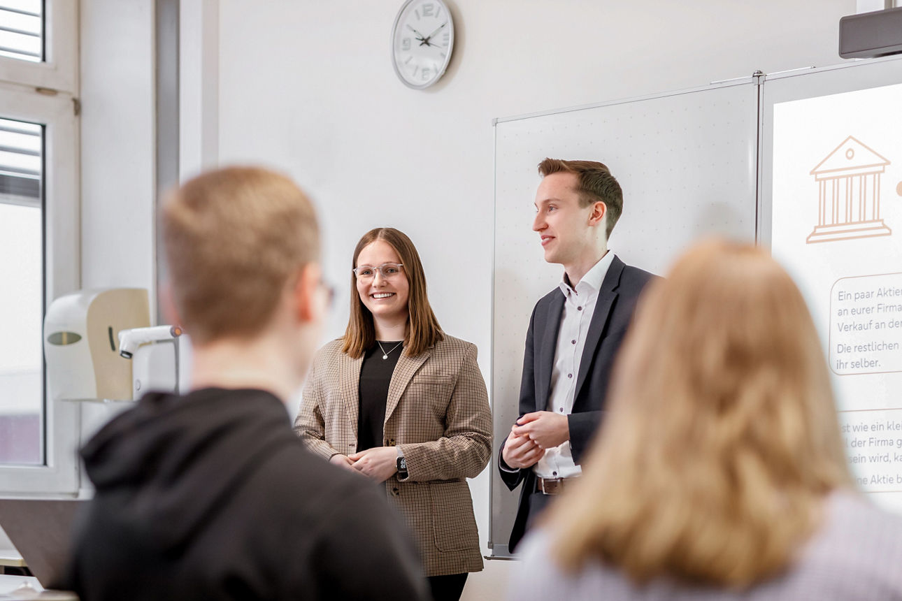 Zwei Personen stehen vor einer Schulklasse und stellen etwas vor