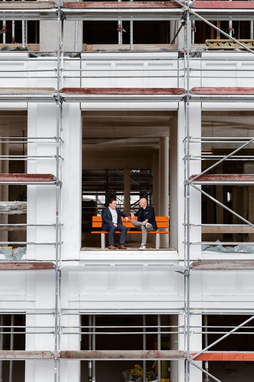 Zwei Personen sitzen auf einer orangenen Bank, die sich in einem Rohbau eines Hauses befindet