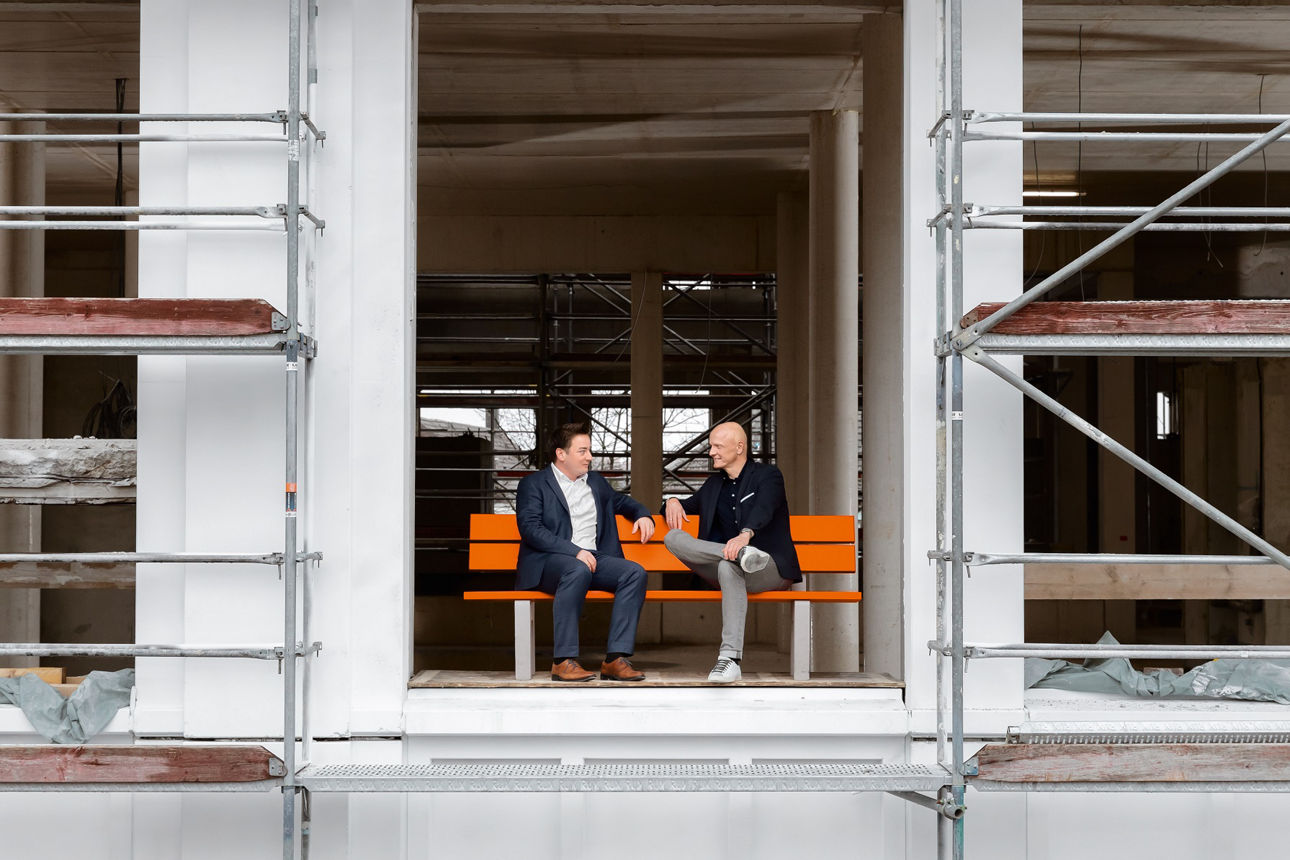 Zwei Personen sitzen auf einer orangenen Bank, die sich in einem Rohbau eines Hauses befindet