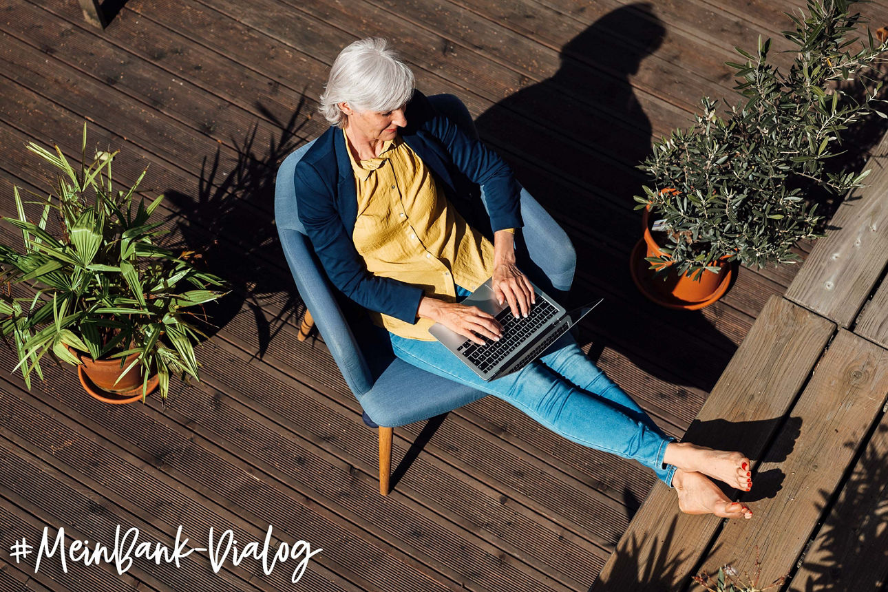 Person sitzt auf einer Terrasse und arbeitet mit einem Laptop; im Bild steht „#MeinBank-Dialog“.
